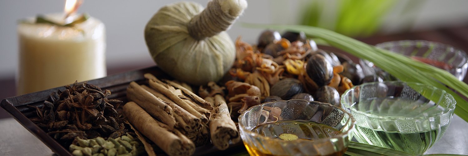 Foto de condimentos ayurvédicos, com raízes, sementes, castanhas óleos, um pinda e uma vela ao fundo.